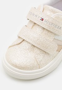 Beige tenisky s texturovaným, lesklým povrchem. Mají dva suché zipy a bílou gumovou podrážku. Logo umístěné na pásku.