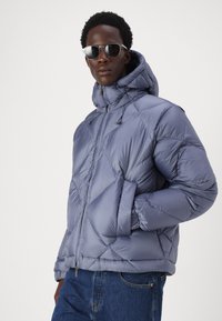 Chaqueta tipo puffer en azul claro con un diseño acolchado, capucha ajustable, cremallera frontal y bolsillos laterales, fabricada en material sintético ligero.