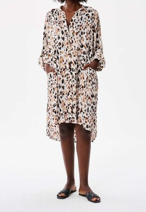 Frau trägt ein locker sitzendes, knielanges Kleid mit langen Ärmeln und Leopardenmuster, das Taschen hat, sowie schwarze flache Sandalen mit offenen Zehen.