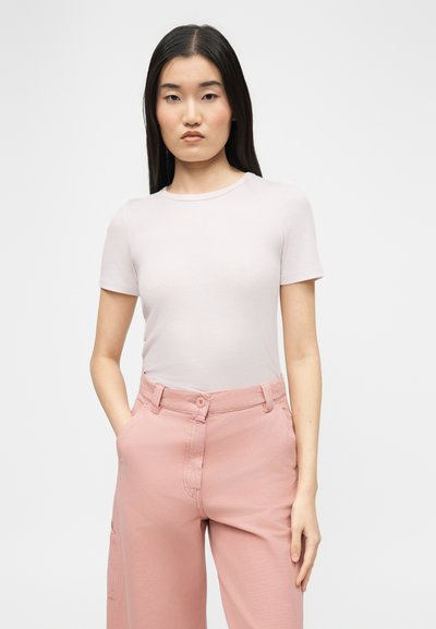 Νεαρή γυναίκα με μακριά σκουρόχρωμα μαλλιά, φορώντας ένα ανοιχτό μπεζ κοντομάνικο πουκάμισο και ροζ ψηλόμεσα παντελόνια, με τα χέρια στις τσέπες, σε ουδέτερο φόντο.