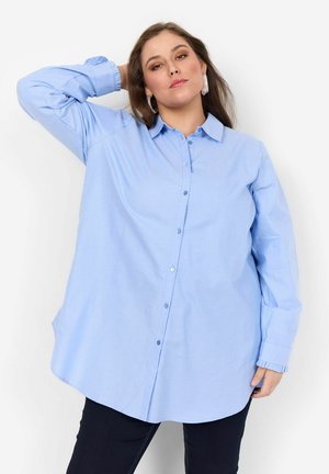 Camisa de botonadura azul claro, hecha de algodón, con un corte relajado, mangas largas y puños con volantes. Textura suave y diseño de cuello clásico.