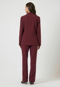 Vrouw staand met de rug naar de camera, gekleed in een donkerrood op maat gemaakt pak met rechte broek en zwarte hakken, tegen een effen lichte achtergrond.