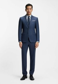 Man staat tegen een witte achtergrond, gekleed in een blauw pak, een overhemd met patroon, een marineblauwe stropdas met stippen en zwarte nette schoenen, handen ontspannen langs het lichaam.