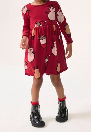 Robe rouge à manches longues avec motifs de bonhomme de neige et de renne, tissu côtelé, jupe évasée et ourlet festonné. Associée à des bottes noires à fermeture éclair.