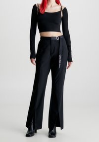 Black long-sleeve crop top with shoulder straps and black trousers featuring side slits and a belt with a logo detail on the side.