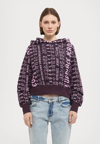 Felpa corta viola con motivi grafici di testo rosa, dotata di cappuccio con cordino e polsini a costina, abbinata a jeans in denim azzurro chiaro.