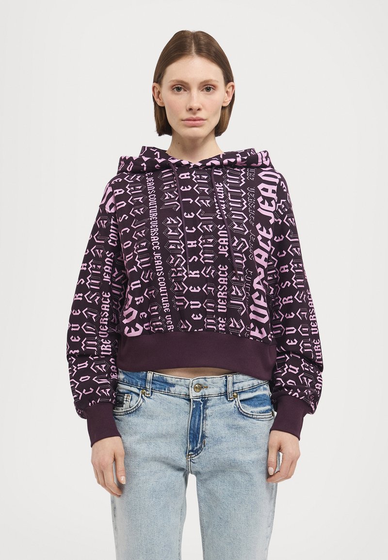 Felpa corta viola con motivi grafici di testo rosa, dotata di cappuccio con cordino e polsini a costina, abbinata a jeans in denim azzurro chiaro.