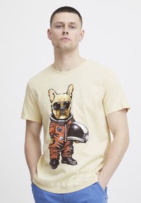 Lysegul t-shirt med en trykt illustration af en hund i en orange rumdragt, der bærer solbriller og holder en hjelm.