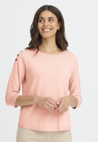 Haut à manches longues rose clair en matière douce, avec des boutons décoratifs sur les épaules et des poignets côtelés. Encolure ronde simple.