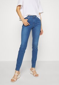 Osoba ve modrých úzkých džínách, bílém krátkém tričku a bílých klínových sandálech, stojící na pozadí z čistě bílé barvy.