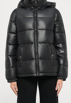 Doudoune matelassée noire brillante avec fermeture éclair sur le devant, poches à pression, poignets élastiques et capuche attachée, portée avec un pantalon noir.