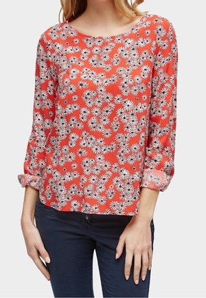 Kobieta w czerwonej bluzce z długim rękawem z białym nadrukiem stokrotek i granatowymi spodniami, rękawy podwinięte do przedramion.