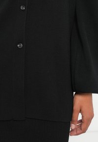 Μαύρο πλεκτό cardigan με ραβδωτή υφή, διαθέτει μεγάλα κουμπιά και χαλαρή εφαρμογή. Περιλαμβάνει μακριά μανίκια και ίσια τελείωση.