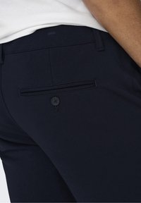 Primer plano de una persona que lleva pantalones azul marino oscuros con un bolsillo trasero abotonado y una camisa blanca metida por dentro.