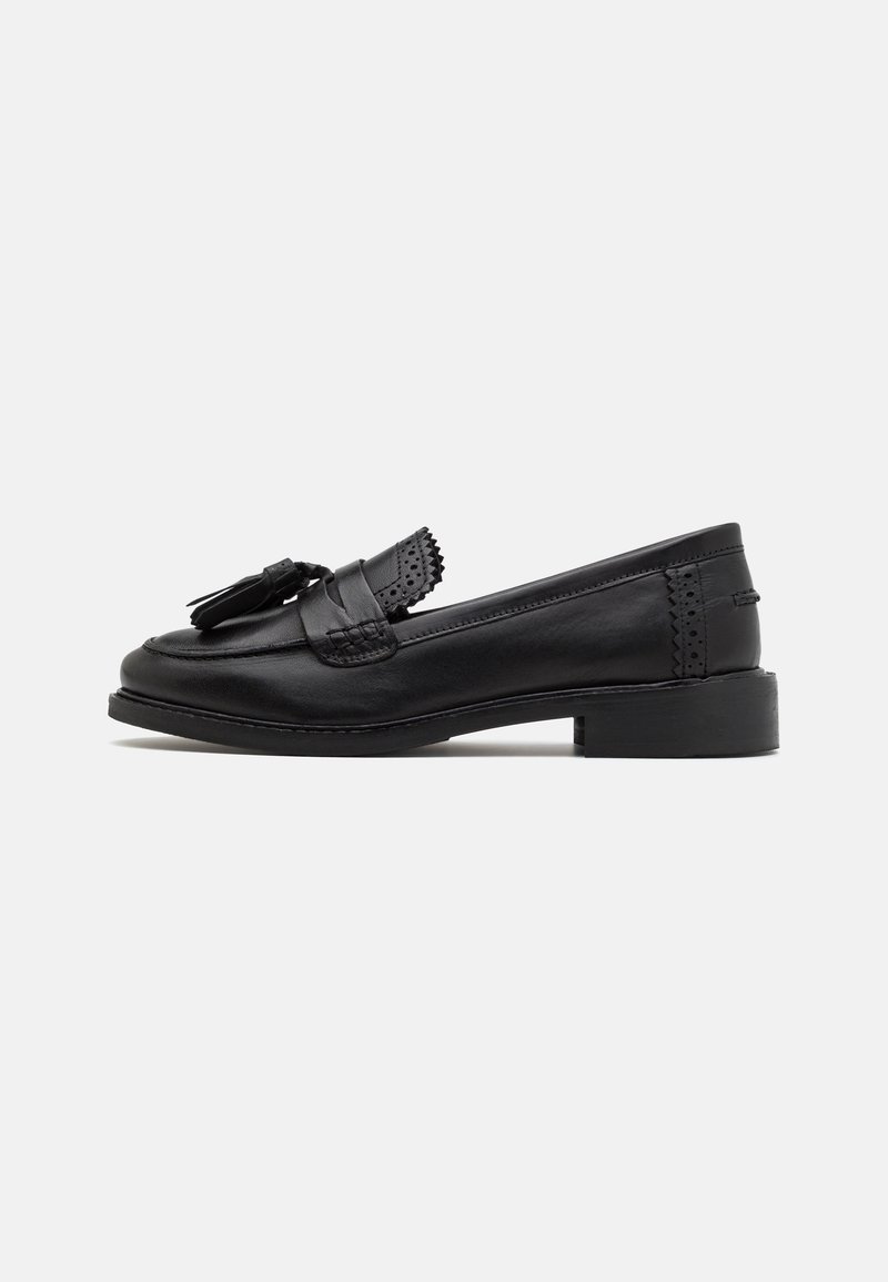 Mocassin à pampilles en cuir noir avec petit talon carré et bordure décorative perforée sur fond blanc uni.