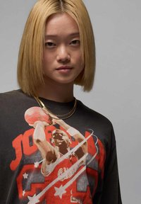 Jeune personne avec des cheveux blonds raides portant un T-shirt noir affichant un joueur de basketball en action et des colliers en chaîne dorés.