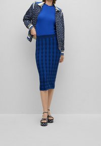 Veste bleue à motifs avec poignets rayés, associée à un haut bleu côtelé et une jupe midi bleue texturée. Sandales à plateforme avec des accents à carreaux.