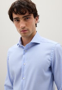 Joven con cabello oscuro que lleva una camisa de botones de rayas verticales en azul claro y blanco, contra un fondo liso.
