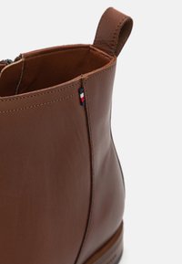 Hnědá kožená kotníková bota s hladkou texturou, bočním zipem a malou vyšívanou vlajkou na lemu.