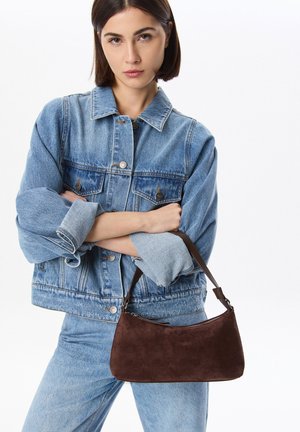 Sac à main en daim marron de forme courbée, avec une bandoulière en cuir courte et une fermeture éclair, associé à une veste en denim et un jean.