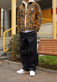 Hnědá džínová bunda s vzorem, bílé mikině s kapucí, černých volných kalhotách a bílých teniskách Nike. Bunda má modré a černé vzory.