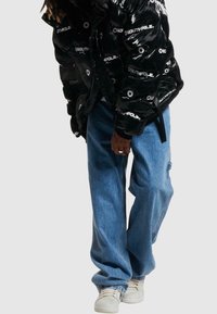 Giubbotto piumino nero con finitura lucida, caratterizzato da stampe di logo bianche; abbinato a jeans baggy blu chiaro e sneakers bianche.