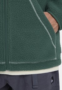 Grüne Fleecejacke mit einem Reißverschluss, ausgestattet mit weißen Nähtelementen und zwei seitlichen Taschen. Getragen über einer hellen Unterlage.