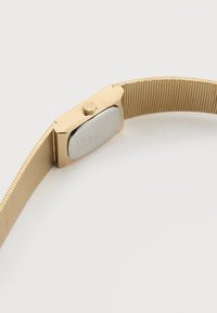 Ρολόι από μέταλλο σε χρυσή απόχρωση με ορθογώνια θήκη και εύκαμπτη συνδεδεμένη μπάντα, εμφανίζεται από πίσω πάνω σε λευκή επιφάνεια.