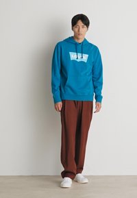 Turquoise hoodie met een wit logo, gecombineerd met roestkleurige wijde broek en witte sneakers. Zachte stof en ontspannen pasvorm.