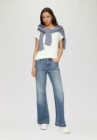 Gestreiftes weißes T-Shirt mit einem lavendelfarbenen Pullover, der um die Schultern gebunden ist, kombiniert mit hellblauen, weiten Jeans und weißen Sneakers.