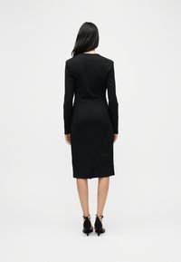 Γυναίκα με μακριά σκούρα μαλλιά, φορώντας εφαρμοστό μαύρο φόρεμα με μακρύ μανίκι και μήκος μέχρι τη μέση της γάμπας και μαύρα γόνατα με λουράκι στον αστράγαλο, στέκεται γυρισμένη προς τα πίσω σε λευκό φόντο.