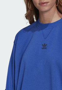 Felpa oversize blu in tessuto morbido con scollo rotondo, caratterizzata da un piccolo logo nero Adidas sul petto e dettagli di cuciture visibili.