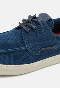 Pantofii de barcă din pânză albastru marin pentru bărbați, dotat cu șireturi gri, talpă din cauciuc și detalii cusute. Design curat cu orificii rotunde.
