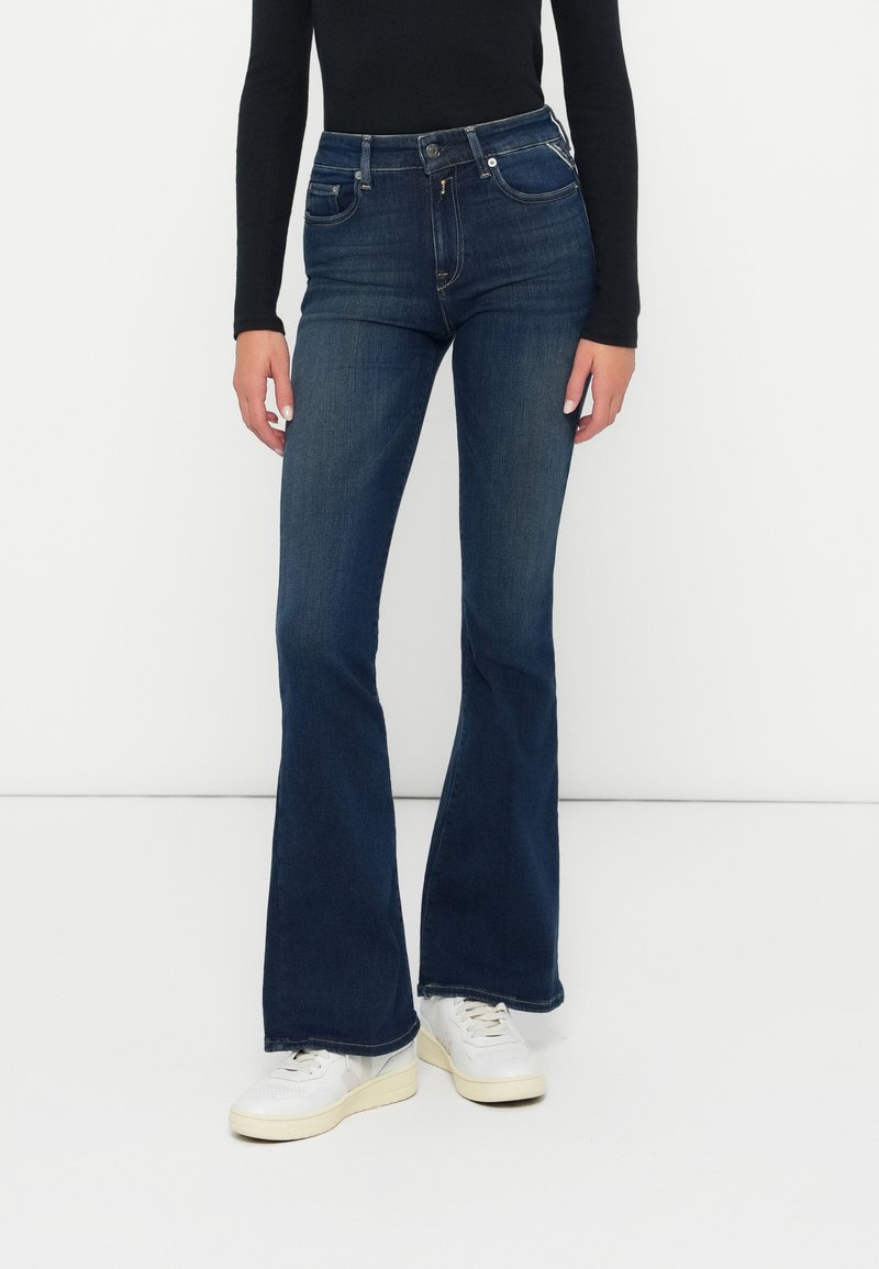 Pantalones vaqueros de denim de corte acampanado en azul oscuro, con una cintura alta, diseño de cinco bolsillos y cierre de botón. Combinados con zapatillas blancas.
