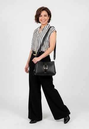 Femme souriante portant un pantalon noir à jambes larges, un chemisier rayé à manches courtes, une ceinture noire, un sac à épaule noir et des chaussures noires pointues.