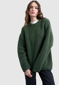 Femme aux longs cheveux ondulés portant un pull vert tricoté épais par-dessus une chemise blanche et un pantalon foncé, debout devant un fond uni.