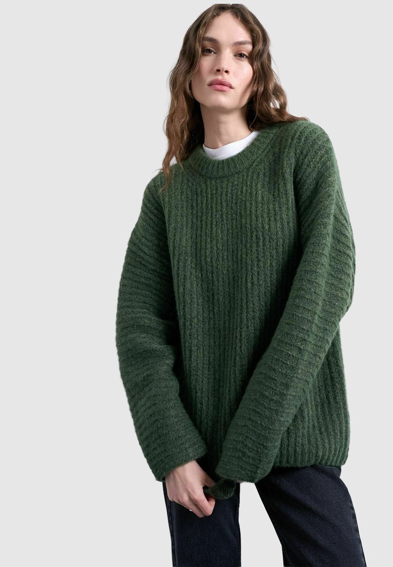 Femme aux longs cheveux ondulés portant un pull vert tricoté épais par-dessus une chemise blanche et un pantalon foncé, debout devant un fond uni.