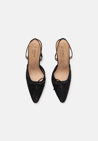 Zapatos de tacón negro con punta afilada y diseño slingback, que cuentan con textura de encaje, pequeños lazos en la parte frontal y una planta en tono nude con marca.