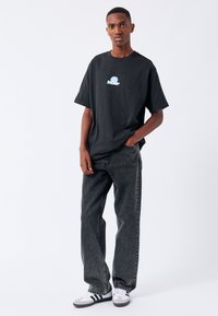Maglietta nera in cotone con grafica blu sul petto, abbinata a jeans grigio scuro. Il modello indossa sneakers nere e bianche.