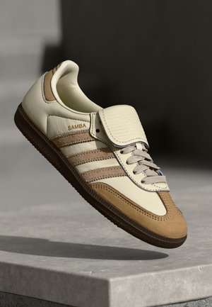 Beige en bruine Adidas Samba sneaker met klittenbandsluiting en veters, getoond in de lucht boven een betonnen oppervlak.
