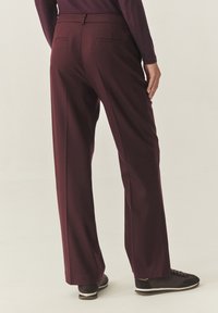 Bordeauxrode broek met een rechte pijp, voorzien van achterzakken en een gladde textuur, gecombineerd met donkere sneakers op een lichte achtergrond.