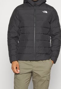 Musta puffatakki, jossa on vetoketjullinen etuosa, huppu, tikattu pinta ja North Face -logo vasemmassa rinnassa.