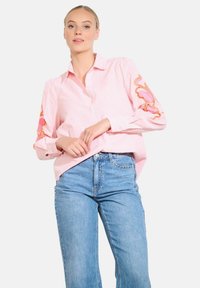 Rožinė dryžuota marškinė su ilgomis rankovėmis, kuriuose siuvinėti gėlių raštai ant rankogalių. Dėvima su aukštu liemeniu mėlynomis džinsomis. Lygi tekstūra.