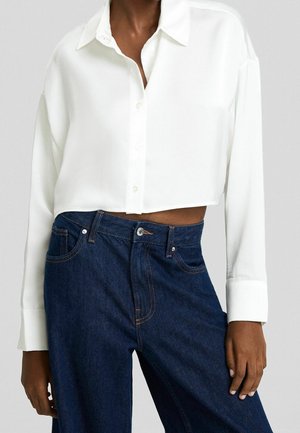 Personne portant une chemise blanche courte à boutons avec des manches longues, associée à un jean bleu foncé taille haute, debout devant un fond uni.