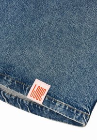 Denimstoff i mellomblått med subtil falming, med et rosa merke preget med "KJÆRLIGHET" i fet oransje skrift.