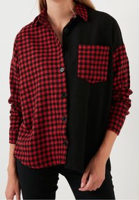 Camisa de franela a cuadros rojos y negros con tela negra en un lado, que presenta un collar, botones y un bolsillo en el pecho. Material texturizado.
