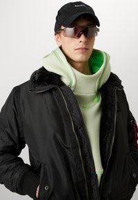 Zwarte bomberjack met een bontkraag, gedragen over een mintgroene hoodie met neonaccenten. Accessoires zijn een zwarte pet en een paar heldere zonnebrillen.