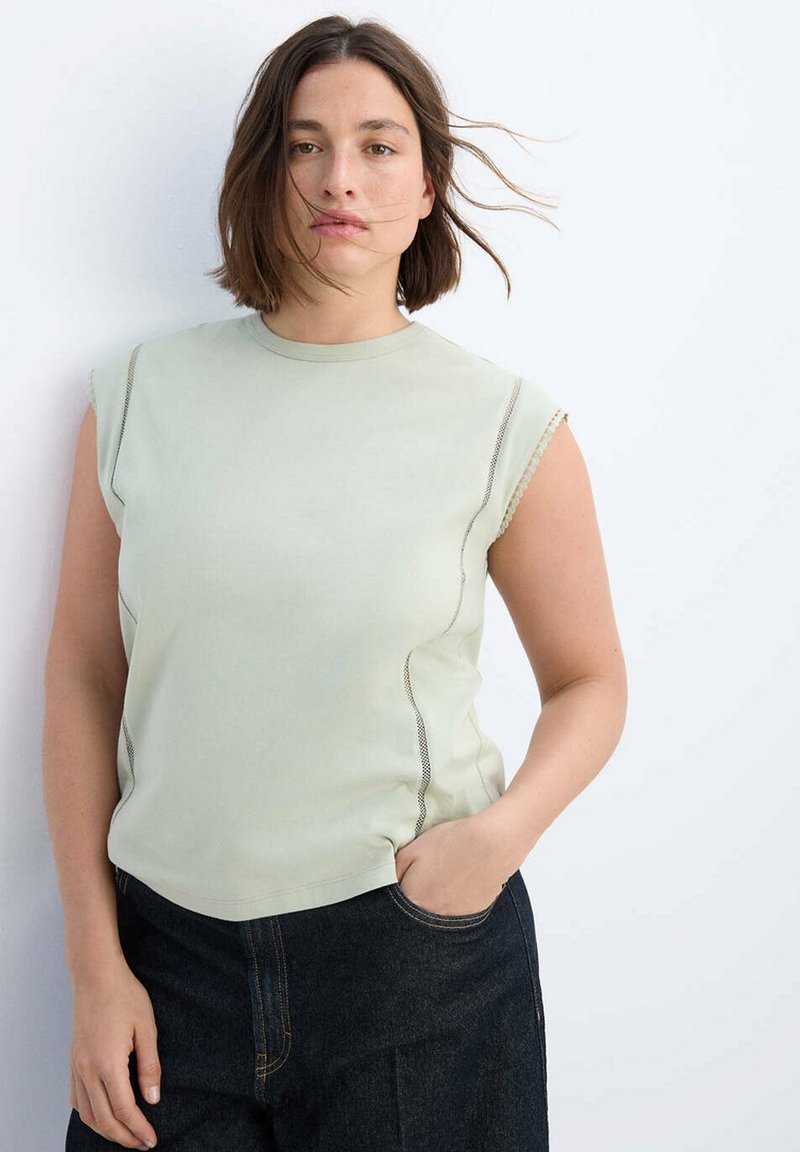 Jeune femme aux cheveux courts bruns portant un haut sans manches vert clair et une jupe en jean foncé, debout, une main dans la poche, sur fond blanc.