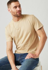 Camiseta de algodón beige con mangas cortas y cuello redondo, combinada con jeans de mezclilla azules. Presenta una cadena dorada y un tatuaje visible en el antebrazo.