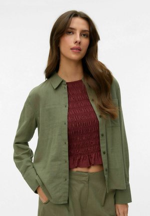 Kobieta z długimi brązowymi włosami, ubrana w otwartą oliwkowozieloną koszulę na teksturowanym bordowym topie odsłaniającym brzuch oraz dopasowanych oliwkowozielonych spodniach, stojąca na białym tle.